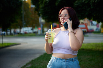 Pretty girl drinking lemonade and talking on phone at sunny day. Young female squinting her eyes because of sunlight. Positive girl drinking and relaxing outside.