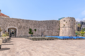 Budva, Montenegro - June 17, 2021: Closed street restaurants near the fortress wall with tower and the Iron Gate of Budva Old Town on a summer morning. Cityscape near a famous tourist resort landmark