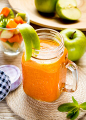 A jar, a glass with orange apple-carrot freshly squeezed juice, a bowl with chopped green apple and carrots..