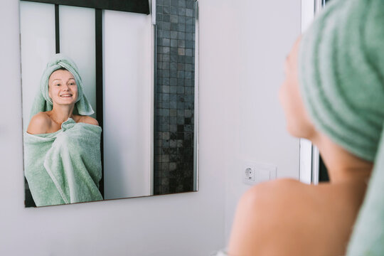 Smiling woman wrapped in towel looking in bathroom mirror