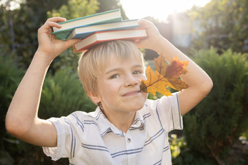 funny blond boy holds an orange autumn leaf in his mouth, and several multi-colored books lie on...