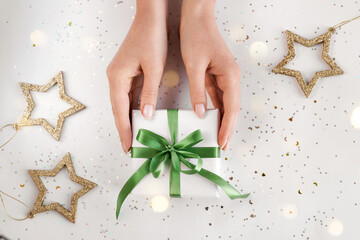 Hands holding Xmas gift package in white wrapping paper with green ribbon on festive background. Zero waste pack Christmas present packaging. Happy holidays concept. Flat lay, top view