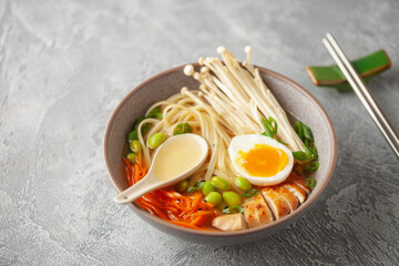 Asian ramen noodles with chicken slice, enoki mushrooms, carrots, edamame beans and egg in a  bowl.