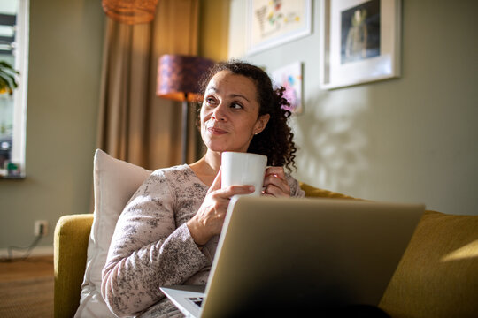 Young mixed woman enjoying a cup of coffee on the couch and using a laptop at home