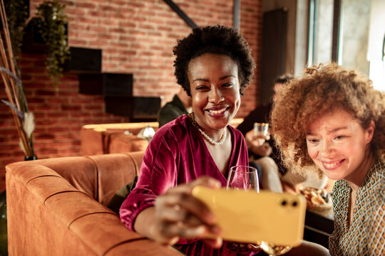 Two young and diverse female friends taking a selfie with their wine on a smartphone at home