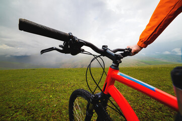 First person view of the handlebars and frame of a bicycle, in the background there are mountains in a fog of clouds. mountain bike on a green meadow on a hill