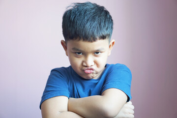 A 6-year-old Asian boy wearing a blue t-shirt stood with his arms crossed over his chest and an angry face.              