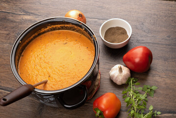 Autumn tomato puree soup in a pan with a ladle and ingredients on a wooden background.