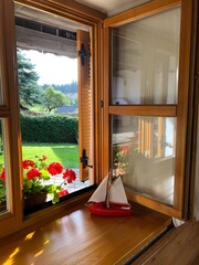 Rotes Spielzeug Segelboot auf einer Fensterbank bei offenem Holzfenster  mit roten Geranien und...