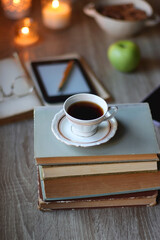 Vintage books, e-reader, pen, reading glasses, phone, cup of tea or coffee, bowl of cookies, green apple and lit candles on the table. Hygge at home, studying atmosphere. Selective focus.