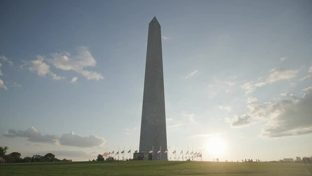 Sunset and flare during golden hour in slow motion of Washington Monument in downtown D.C. by mall, American flags, and people visiting on vacation during a bright summer evening