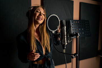 girl singer singing in a recording studio with headphones in front of microphone to create a new...