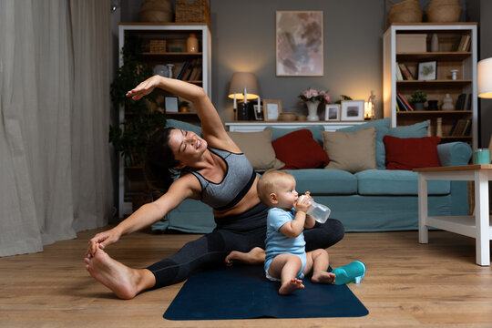 Young happy successful independent single mother is practicing yoga at home on a yoga mat while her baby is carelessly sitting on the floor and playing with a water bottle. Mom stretching muscles