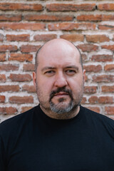 Outdoor close-up of a 45-year-old bald man with a goatee and mustache facing the camera against a brick background