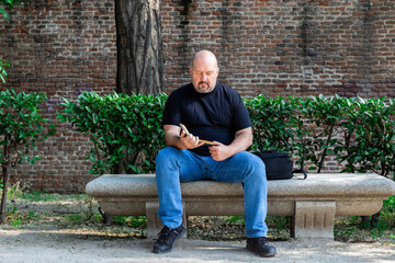 Fototapeta premium Portrait of a bald man in a black T-shirt sitting on an outdoor park bench concentrating on reading a book