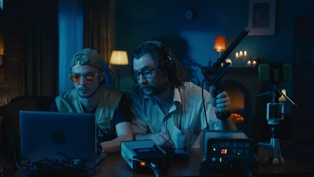 Medium-sized video of two ghost hunters sitting at the table in a dark room trying to capture the activity of an entity that's making the lights flash with an EMF detector.