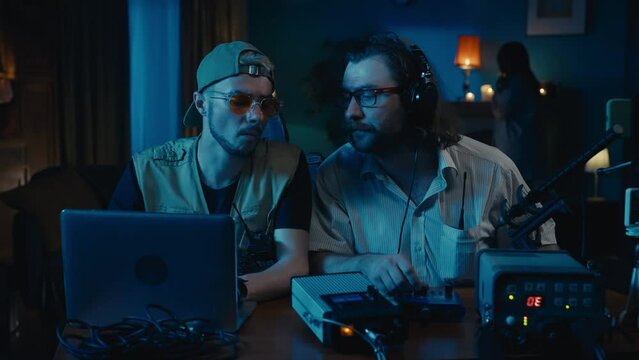 Medium-sized video of two ghost hunters sitting at the table in a dark room trying to capture paranormal activity with an EMF detector. The victim is wandering behind.