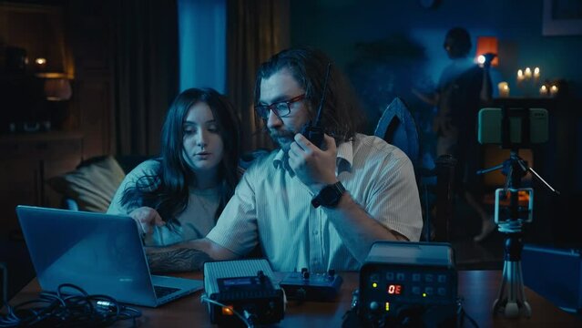 Medium-sized video of a ghost hunter and a victim sitting at the table in a dark room. Another ghost hunter is trying to capture paranormal activity with an EMF detector.