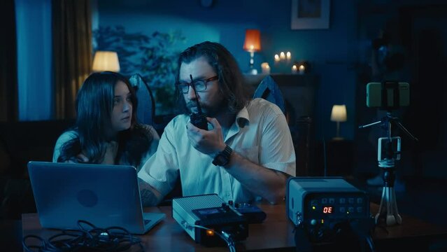 Medium-sized video of a ghost hunter and a victim sitting at the table in a dark room. Another ghost hunter is trying to capture paranormal activity with an EMF detector.