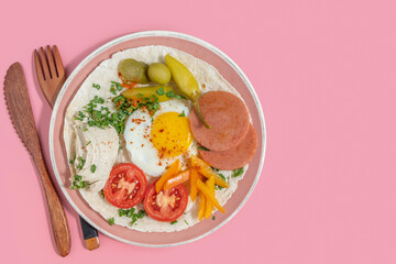 Breakfast plate with sunny side up egg, fresh and pickled vegetables, roasted sausage, creamy hummus on pink background.