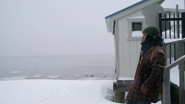 Profile shot of a woman in winter clothes standing near lake with pier on a snowy shore during blizzard, holding hands in pockets, looking at distance. High quality 4k footage