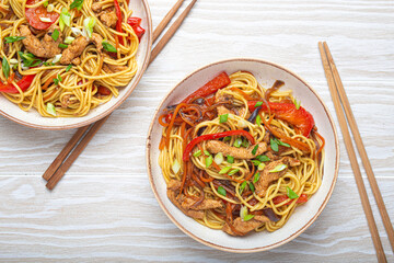 Two bowls with Chow Mein or Lo Mein, traditional Chinese stir fry noodles with meat and vegetables, served with chopsticks top view on rustic white wooden background table.