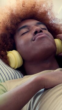 Vertical video. African young man lying on a sofa with eyes closed listening to music with headphones. Relaxation on the sofa 