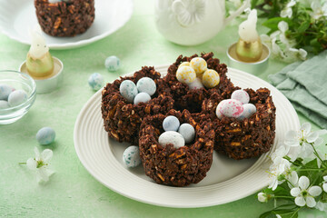 Easter chocolate nest cake with mini chocolate candy eggs with blossoming cherry or apple flowers on green background table. Creative recipe for Easter table with holiday decorations. Top view.