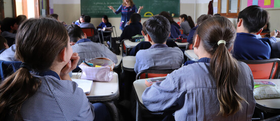 Group of students in a classroom with a teacher explaining a topic.  Boys at their desks dressed in...