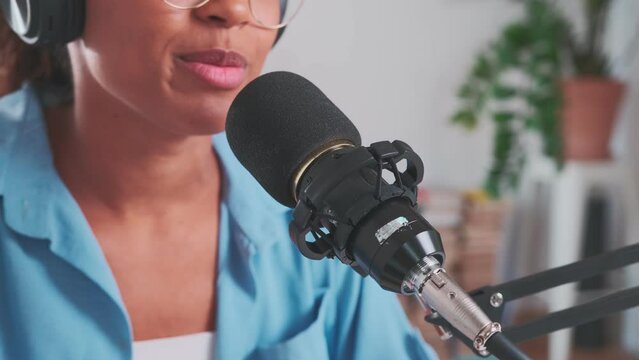 Young cheerful African American woman reads text into microphone mounted on tripod and laughs communicating with radio listeners dressed in blue shirt and glasses sits at table in recording studio.