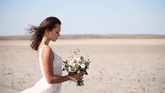 Romantic bride with delicate wedding bouquet at desert landscape. Hair of pretty woman blowing in a wind in nature. Elegance lady with pink, white fresh flowers waiting for groom. Wedding photoshoot.