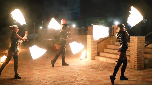 Artists spin burning poles at night fire performance show. Male, female dancers troupe act with torches at the street festival. Fire arts team juggling with flames outdoor in the darkness.