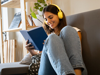 Young woman reading a book happy while listening to music with headphones lying on the sofa in an apartment - obrazy, fototapety, plakaty
