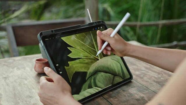 Female artist hands drawing with stylus on tablet at the cafe on tropical island, palm trees. Young designer works remotely open air at exotic location. Pretty woman editing illustration. Close-up.