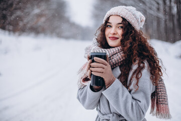 Beautiful girl wearing knitted hat and scarf, holding thermos with hot tea in frosty winter day.