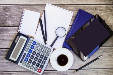 On a wooden background, an economist’s desk, a tablet, a notepad, pens, pencils, a calculator, a magnifying glass, sheets of paper, a cup of coffee.  Office work concept, coffee break, flat top view.