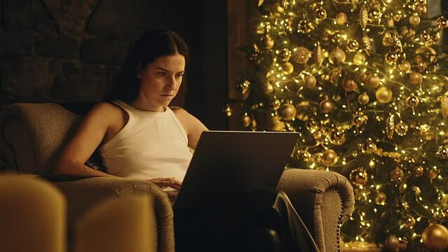 Young beautiful business woman working on a laptop with a colorful Christmas tree in the background. Christmas tree. Cozy Christmas atmosphere. 4K