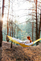 A girl in a hammock in an autumn pine forest looks at the sunset
