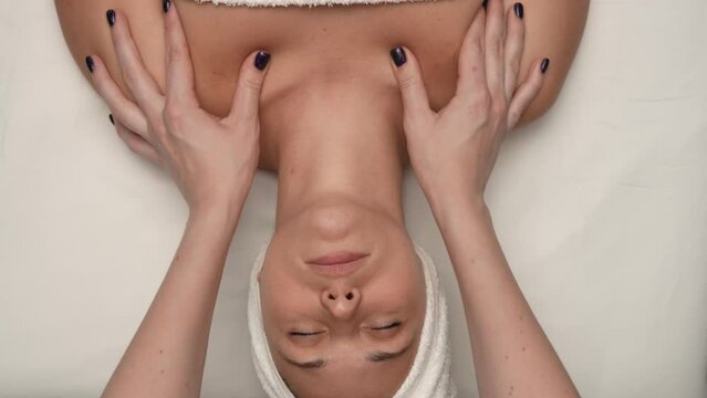 Top view of woman on massage table covered by towel, female therapist massaging her collarbone and neck area. Slow motion.