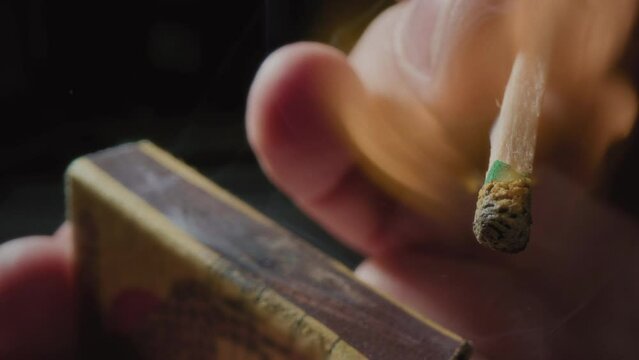 Hands lights a match with a green sulfur head, slow motion. Strikes a match on the side of the box. Smoke and fire appear. The flame ignites. The match lights up, burns and goes out. Super macro shot