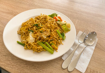 Fried noodle on white ceramic plate with pair of spoon and fork placed on tissue isolated on wooden...