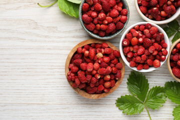 Fresh wild strawberries in bowls and leaves on white wooden table, flat lay. Space for text