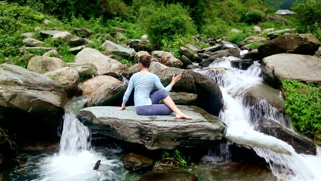 Yoga exercise outdoors - woman doing Ardha matsyendrasana asana - half spinal twist pose at tropical waterfall in Himalayas in India. Vintage retro effect filtered hipster style image.