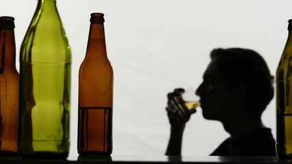 In the close-up shot, there are empty, broken glass bottles full of alcohol. In the background, a strong man holds a glass of alcohol in his hands and examines it. Then he drinks sad and sad