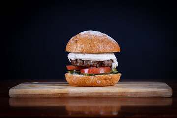 Beef burger with tomato, cucumber, cheese and sauce served on cutting board on a black wooden...