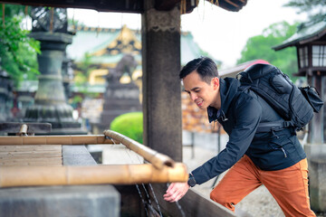 Side view of smiling young Hispanic male tourist backpacker with rucksack touching flowing water in old fountain Kaneiji temple on sunny day in Japan, Tokyo - obrazy, fototapety, plakaty