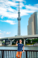 Full body of Hispanic male tourist traveler in casual outfit and sunglasses standing on embankment and taking selfie with smartphone near Tokyo Skytree observation tower under blue sky, Japan