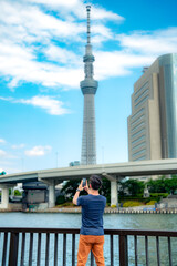 Unrecognizable full body of Hispanic male tourist traveler in casual outfit and sunglasses standing on embankment and using smartphone near Tokyo Skytree observation tower under blue sky, Japan