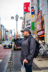 Calm Hispanic male tourist in outerwear and cap with backpack and umbrella walking on city street near tall buildings in Akihabara neighborhood in Tokyo, Japan