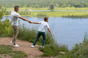 Naklejka na ściany i meble Father and son in white T-shirts walking, chatting on the riverbank on a sunny afternoon. High quality photo